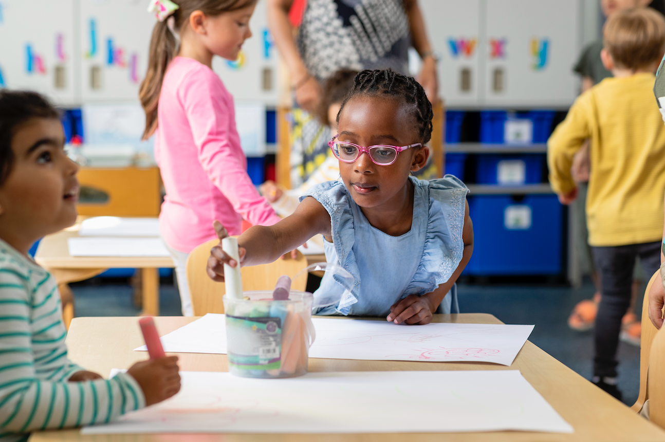 an elementary-age child in classroom reaching for a writing utensil over the desk