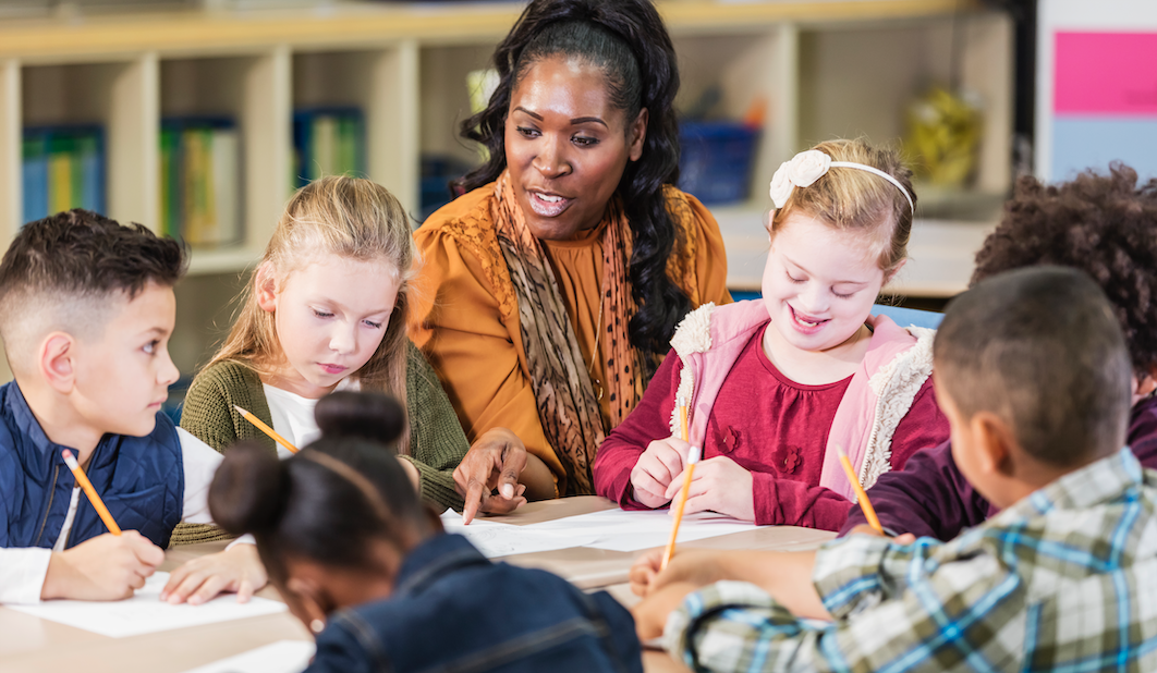 a group of elementary-age children sitting at a table around their teacher who helps them with an assignment